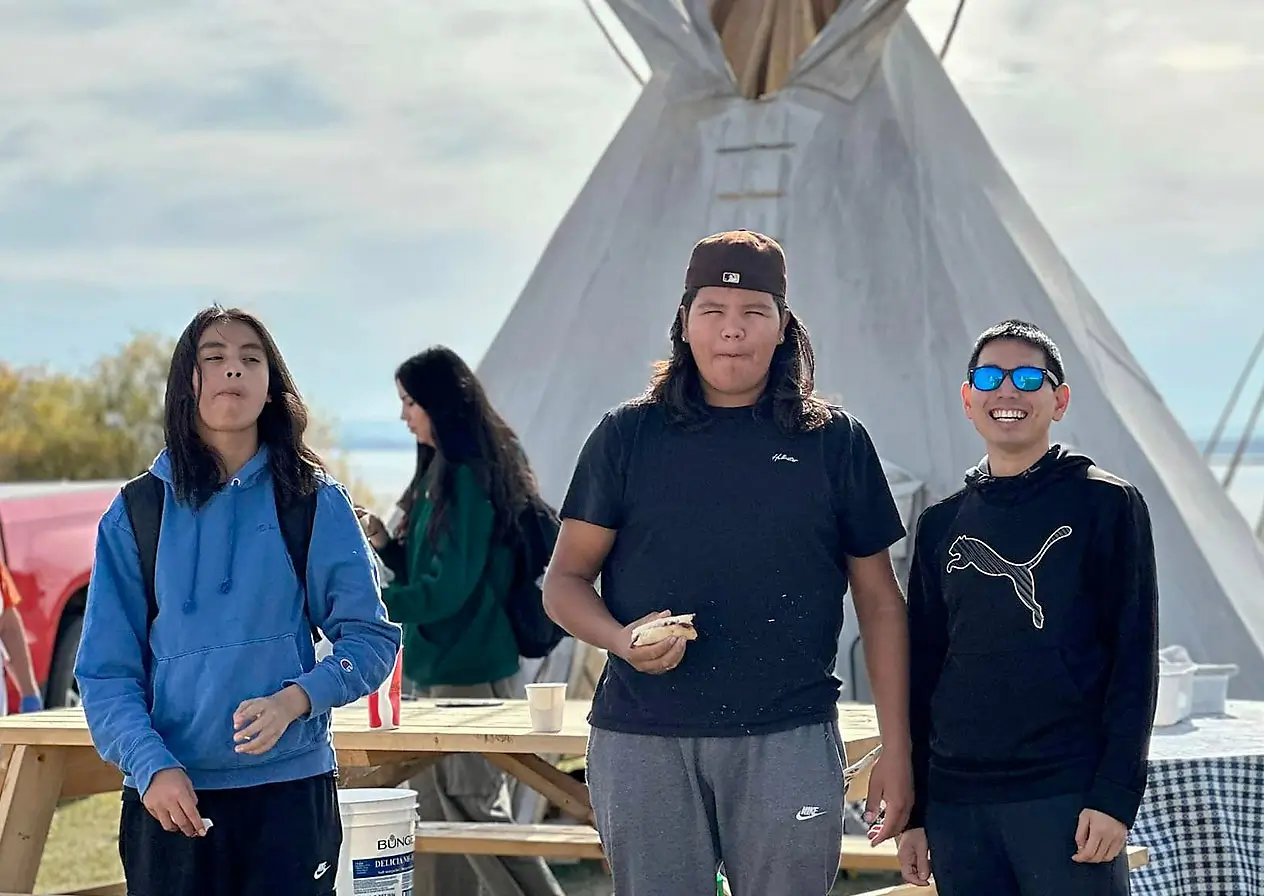 3 youths standing in front of a picnic table and tipi